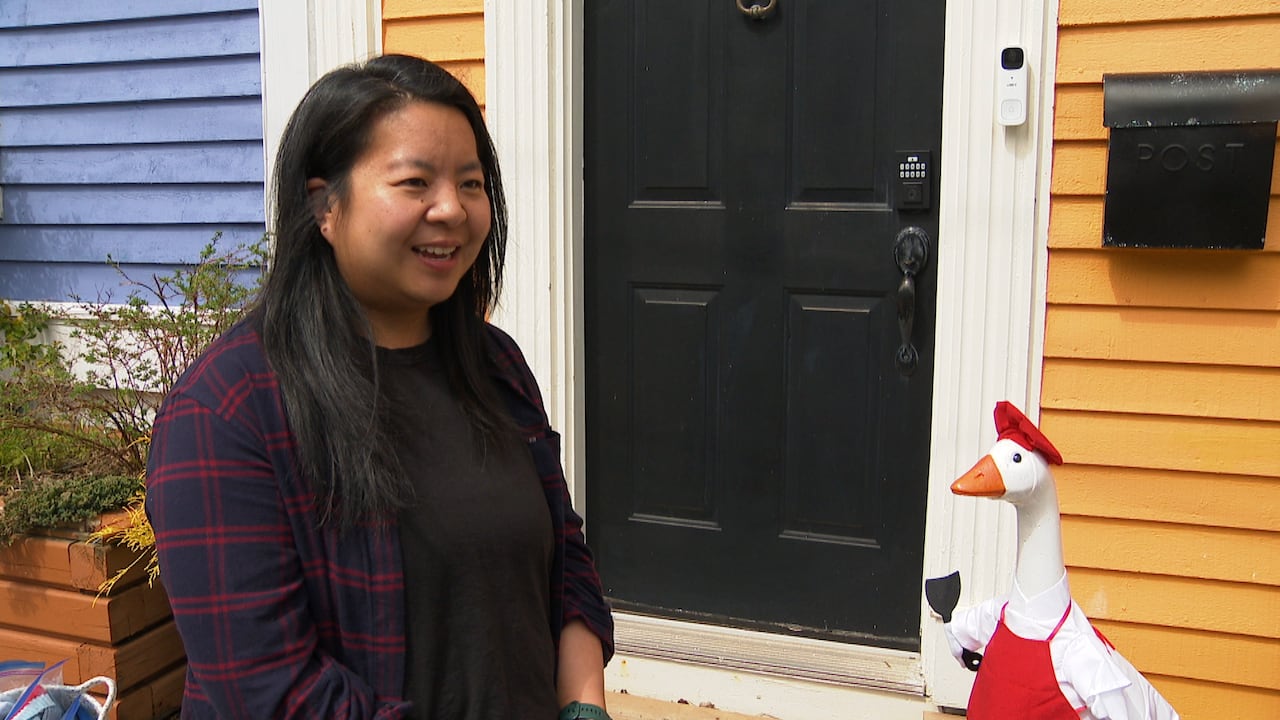 A smiling woman stands on the porch of her home. There's a resin goose sitting on the porch, dressed like a chef with an apron, mini chef hat and a cooking utensil.