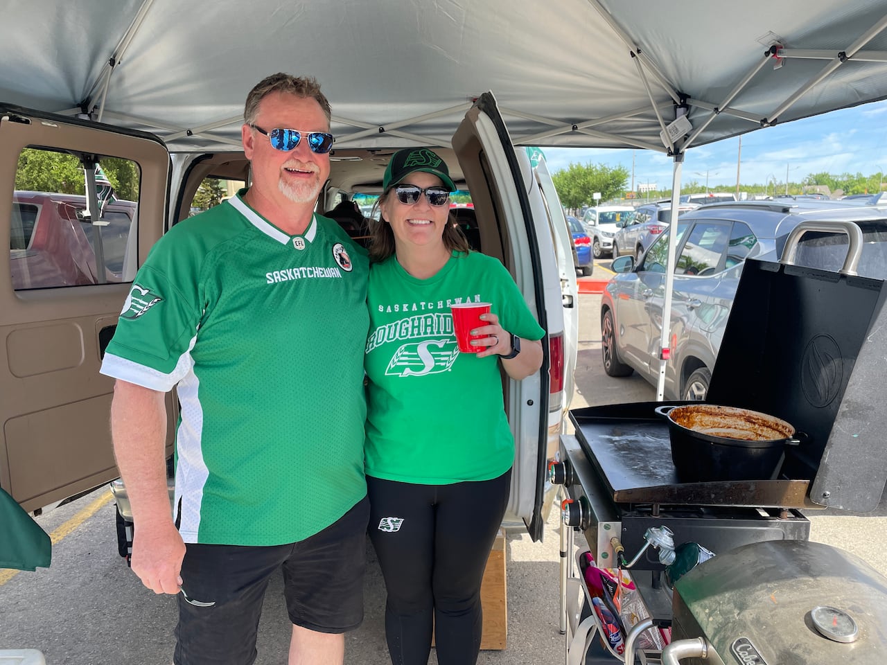 A man and a woman both wearing green Saskatchewan Roughrider shirts stand next to a propane grill with a pot on it.