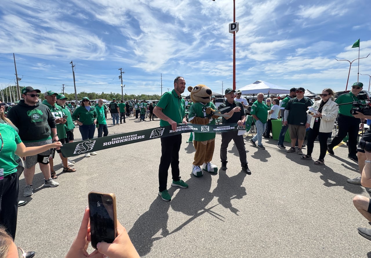 Two men and a gopher mascot stand behind a ribbon that says Saskatchewan Roughriders. The gopher mascot is holding a large pair of scissors to cut the ribbon.