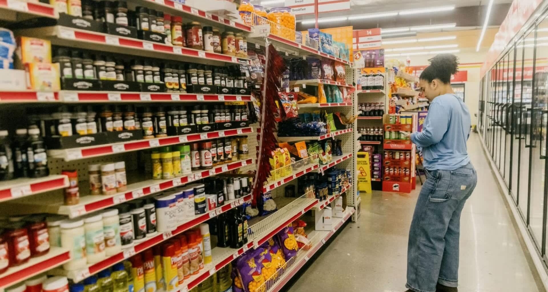 A person stands in a grocery aisle, browsing a variety of spices, sauces, and snacks on the shelves.