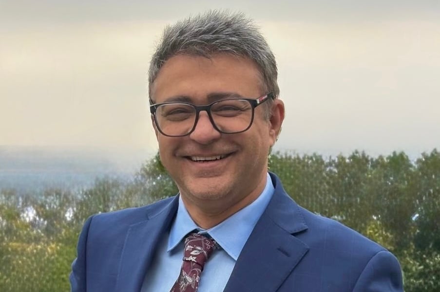 A man in a dark blue blazer and a light blue shirt poses for a photo while standing outside.