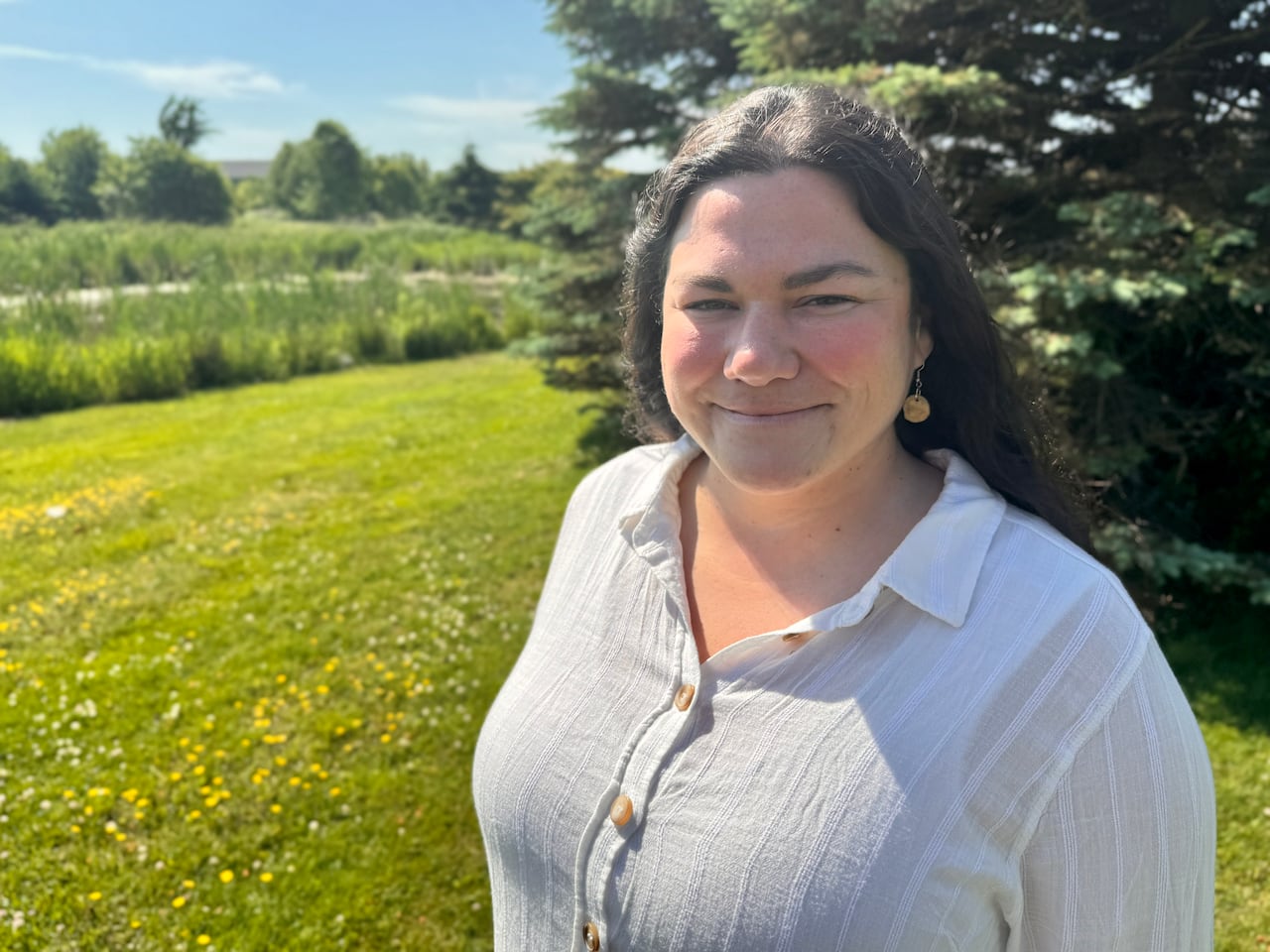 A woman smiles outdoors, wearing a white shirt, against a grassy background with trees and blue sky