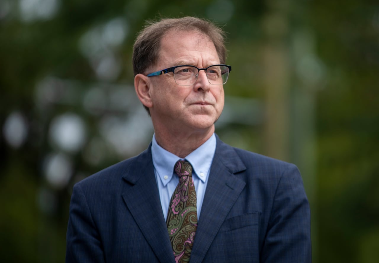 A man wearing glasses and a patterned tie is seen looking off into the distance.