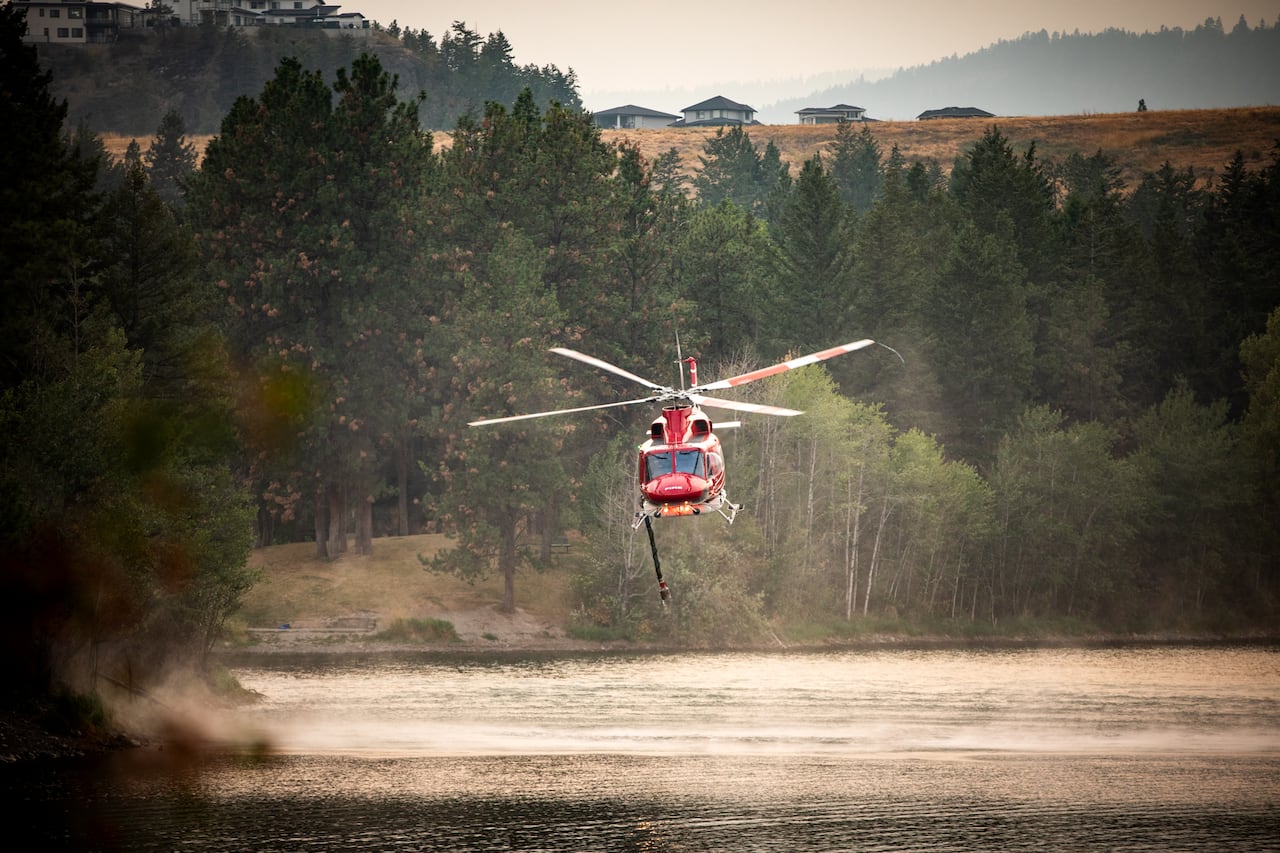 A red helicopter hovering near a lake in a amber-hued forested area.