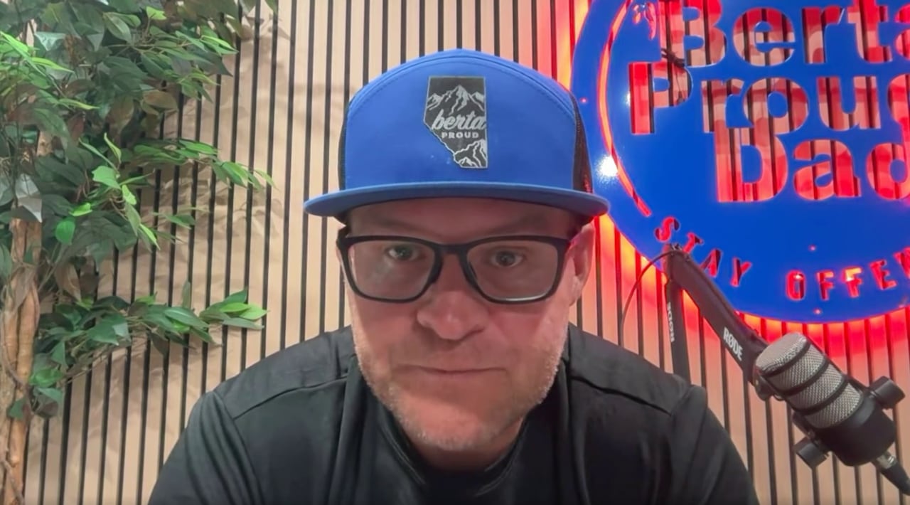 A man wearing a blue ballcap that says 'Berta proud' on it sits in front of a microphone.