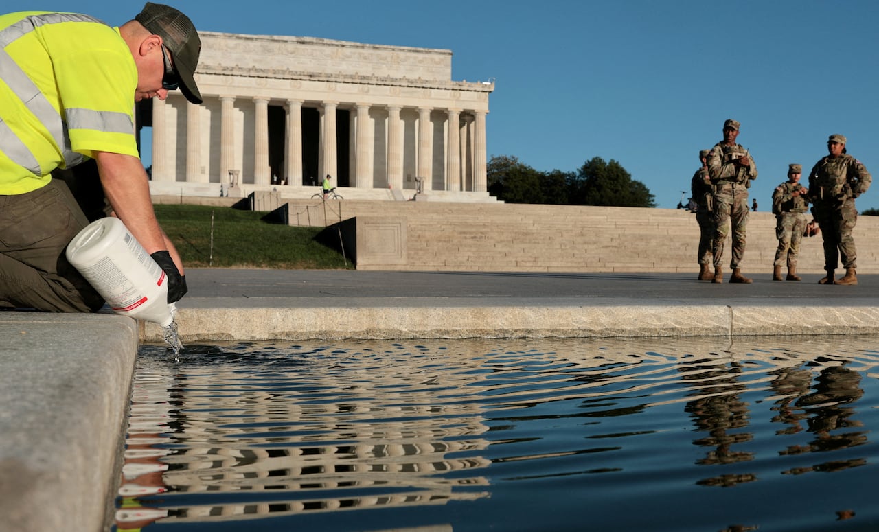A worker wearing a yellow reflective vest pours liquid from a white plastic jug into the pool, with the Lincoln Memorial and four National Guard members visible in the background.