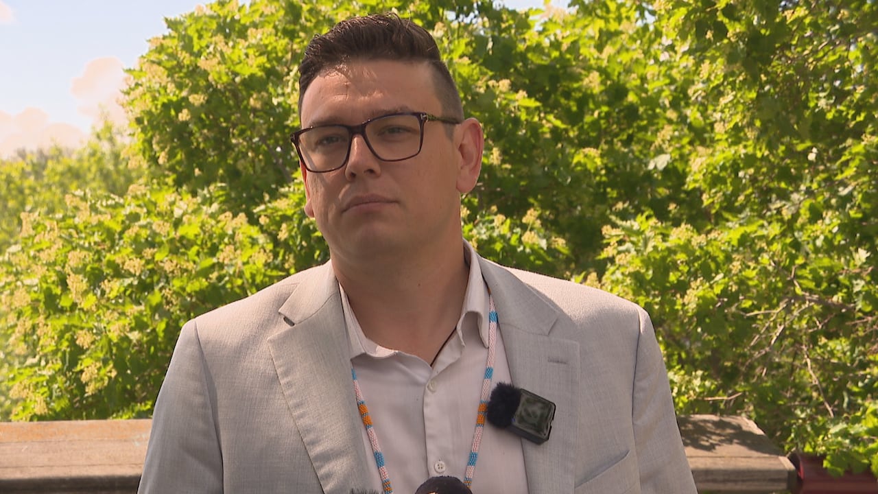 A man in a suit and glasses looking forward with green trees behind him