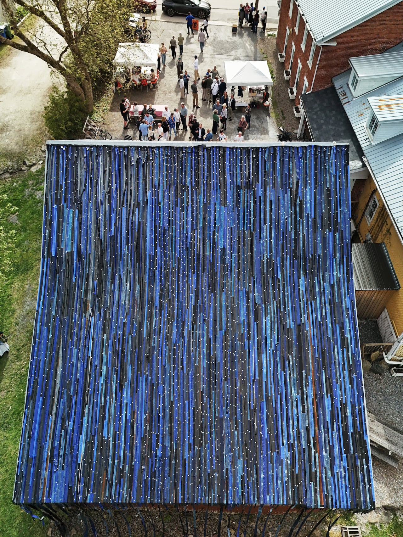 Drone view of a roof covered with a striped and shimmering blue textile artwork. People in a crowd can be seen gathering on the ground below.