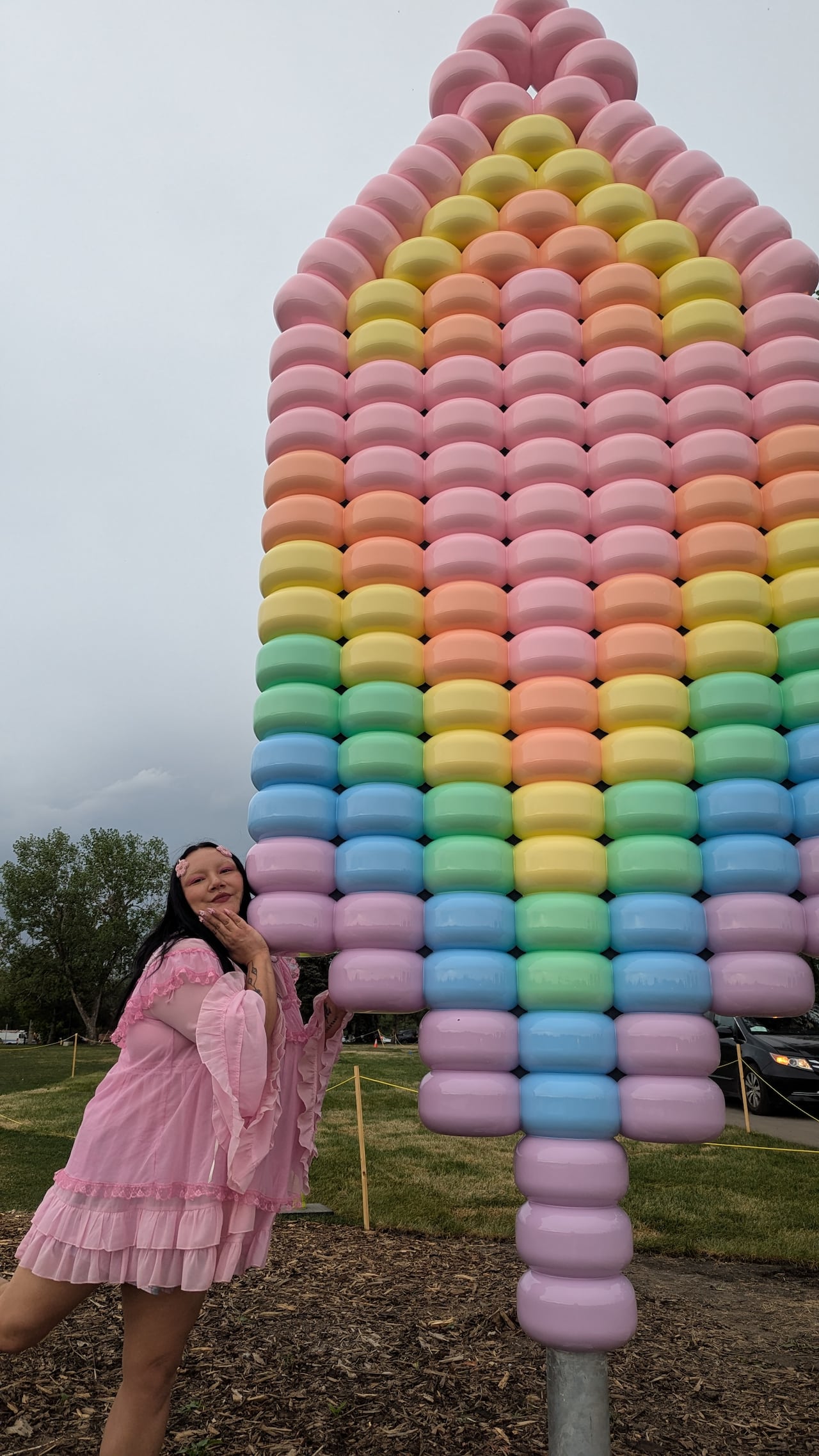 A woman in a pink ruffled mini dress, artist Cheyenne Rain LeGrande, smiles and poses next to a tall beaded sculpture resembling an earring in pastel rainbow colours. It is in a park with green grass and grey skies.