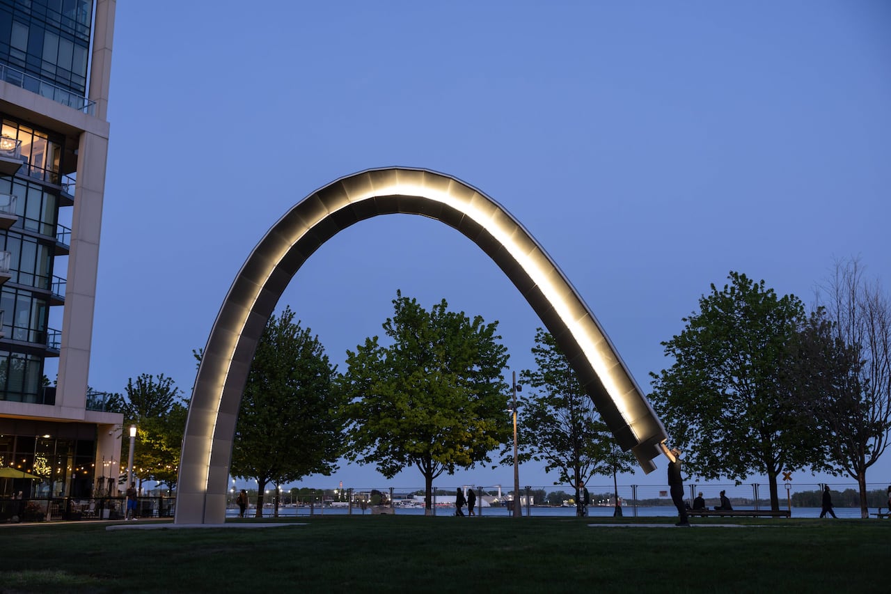 Photo at dusk along Toronto waterfront. A monumental sculpture in the shape of an arch lights up. A man, seen in profile, stands and raises his hands to touch the sculpture, connecting the arc shape to the ground.