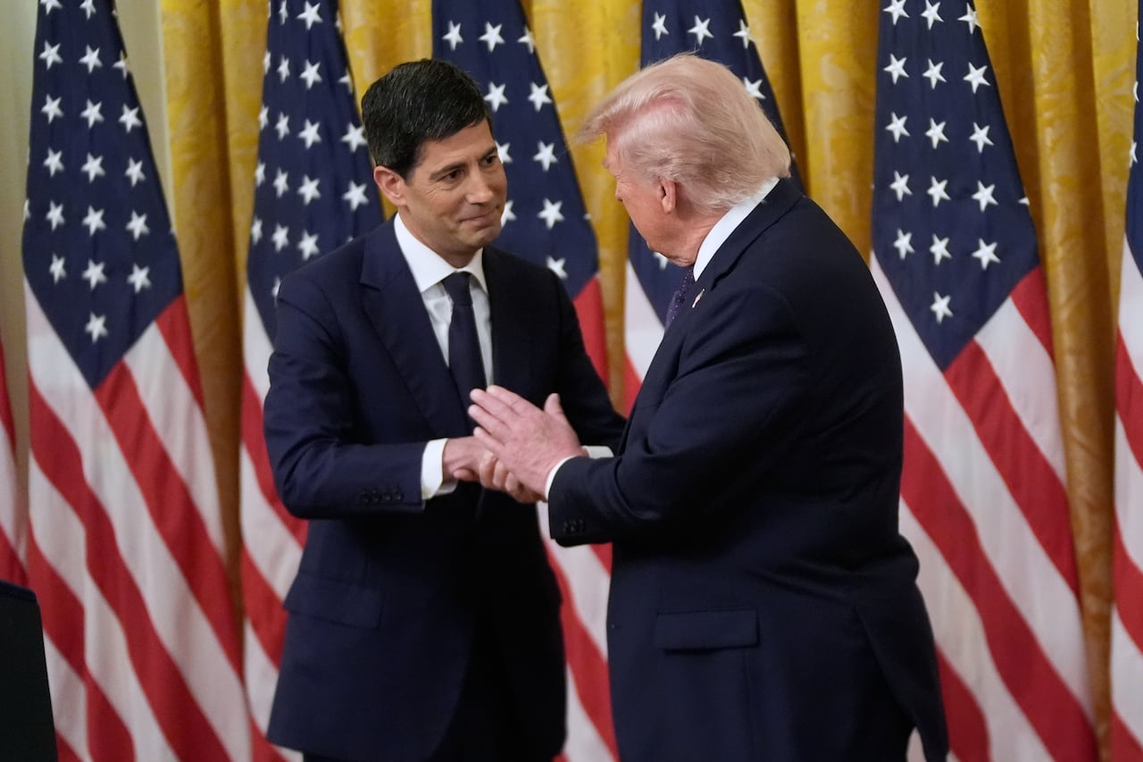 two men in suits shake hands on a stage in front of a row of american flags