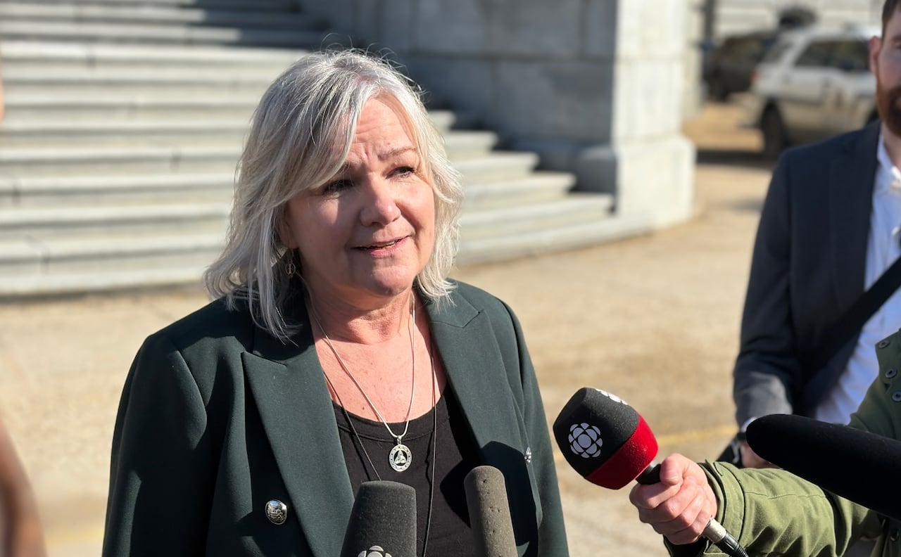 A woman in a forest green blazer stands in front of the legislature building in the sun. Reporters hold microphones up to her.
