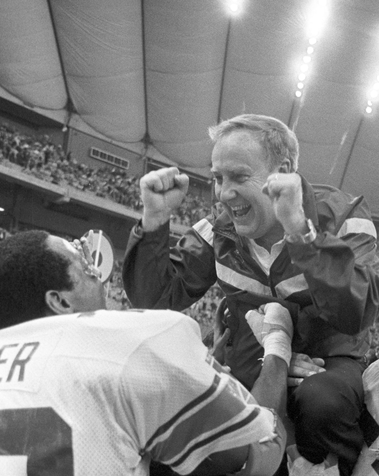 A football coach raises his fists in celebration while being lifted by a player.