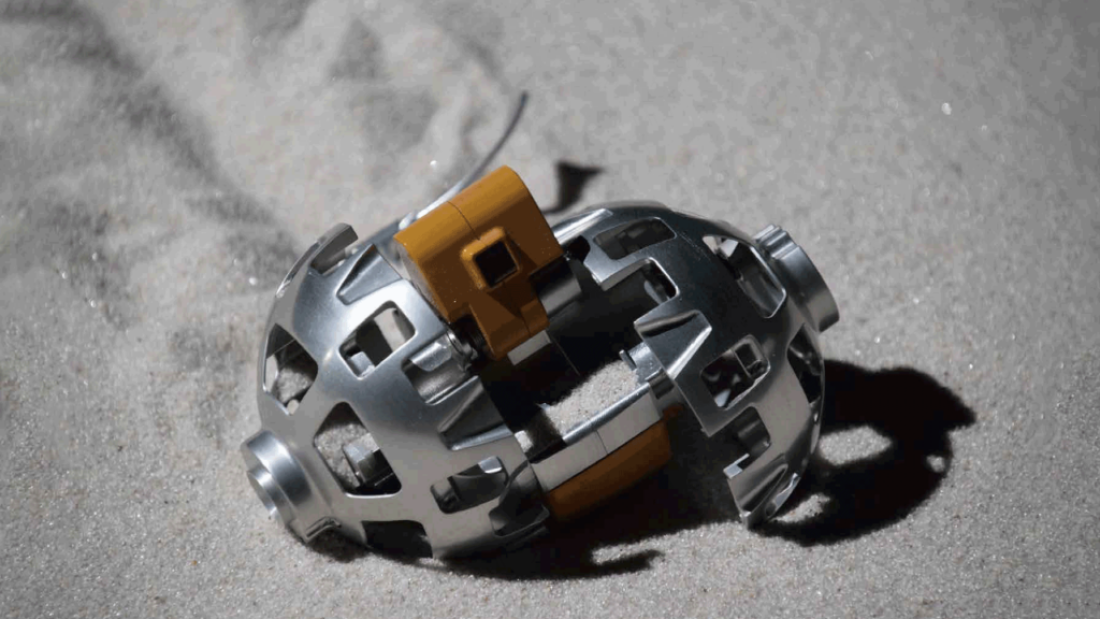 A gray metal cage-like sphere is partially open in the sand revealing a tannish orange internal square.