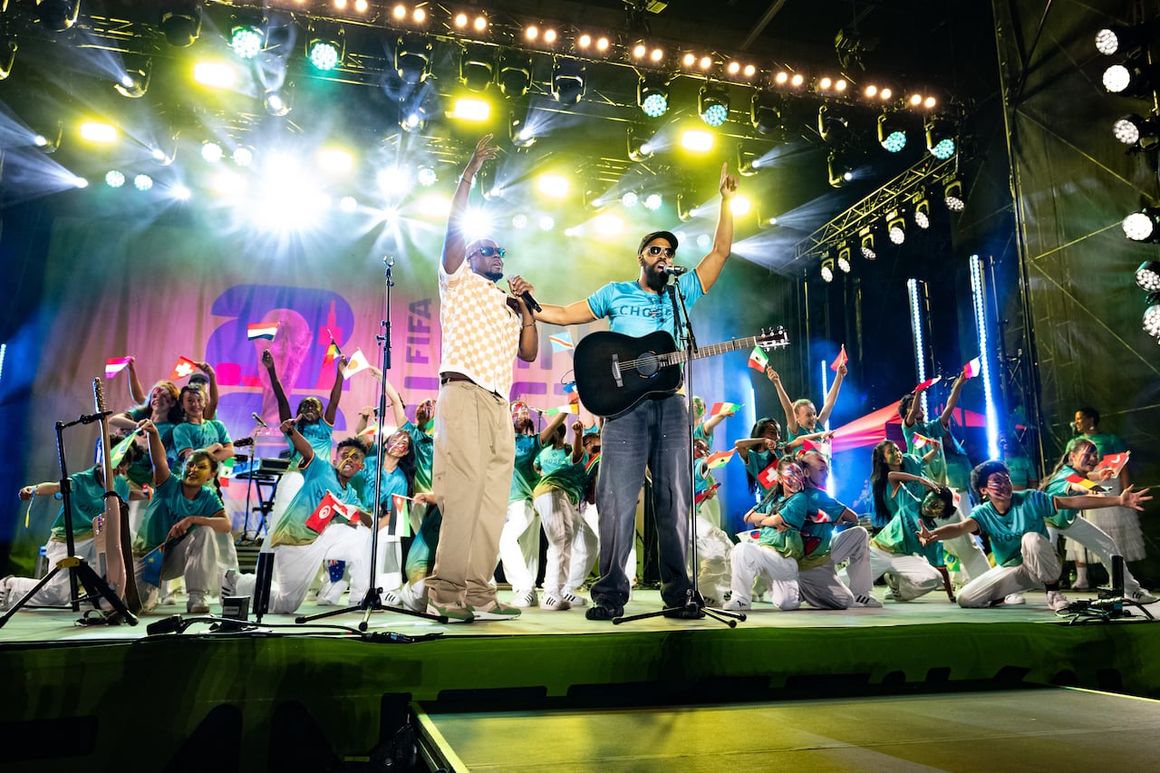 Two singers stand on a stage amid bright lights, surrounded by dancers holding flags of the world.