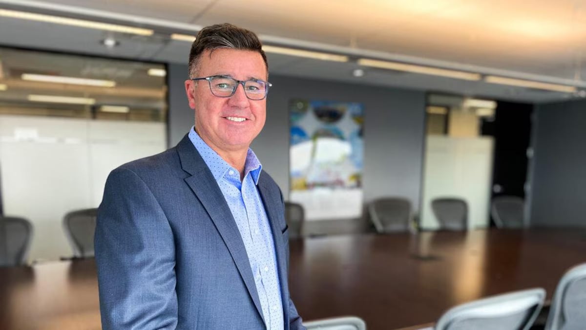 A man in a blue suit shirt and blazer and glasses is standing in a boardroom.