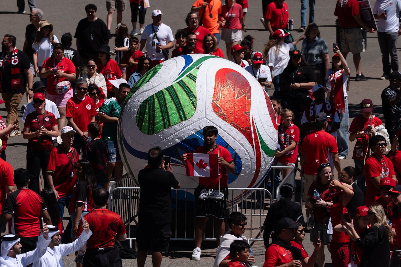 Many fans wearing red wander around a large soccer ball, as one fan poses for a photo holding a Canadian flag.