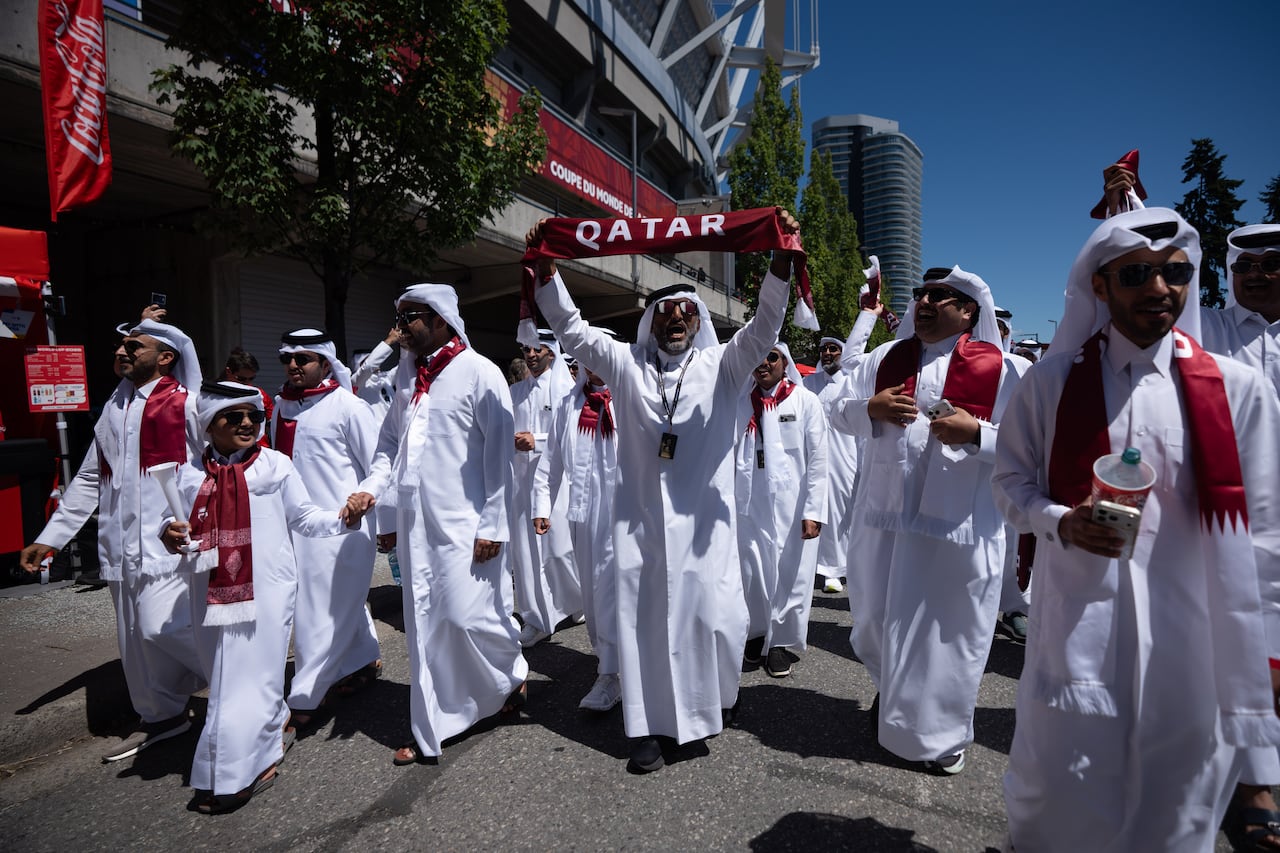 Qatar fans march outside B.C. Place wearing white robes and maroon scarves that say "Qatar." One man in the middle holds up the scarf.