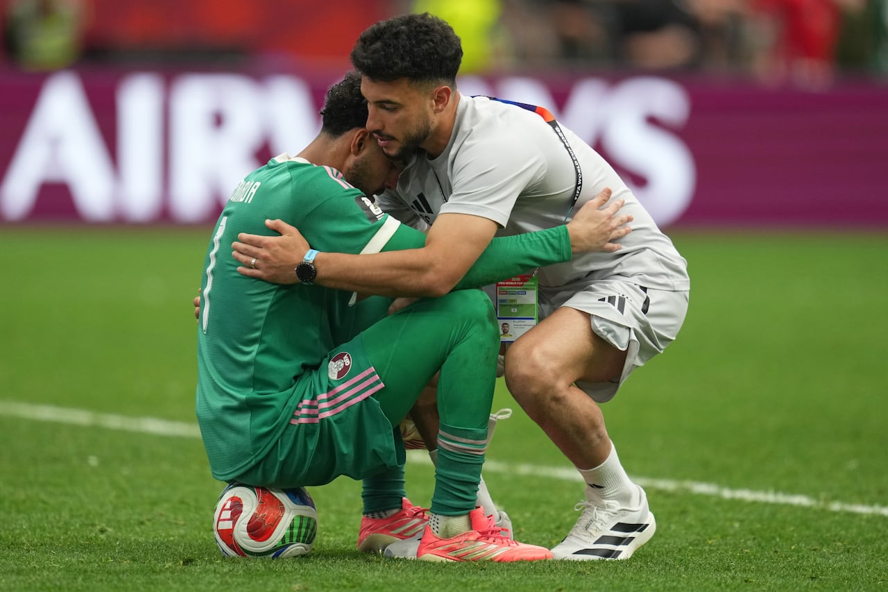 A soccer goalkeeper sitting on a ball embraces a team member on the pitch