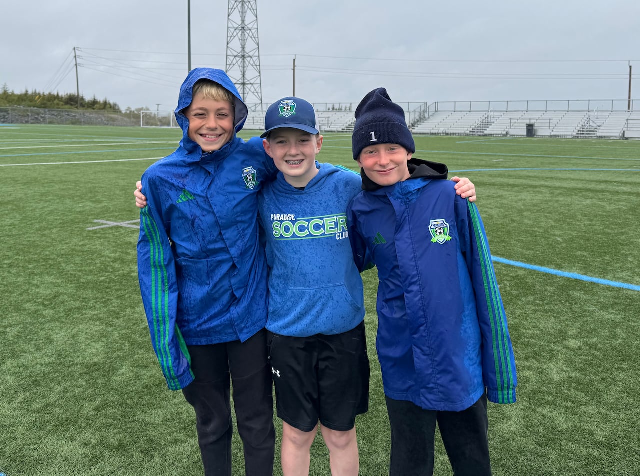 Three boys smiling with their arms around each other standing on a soccer field.