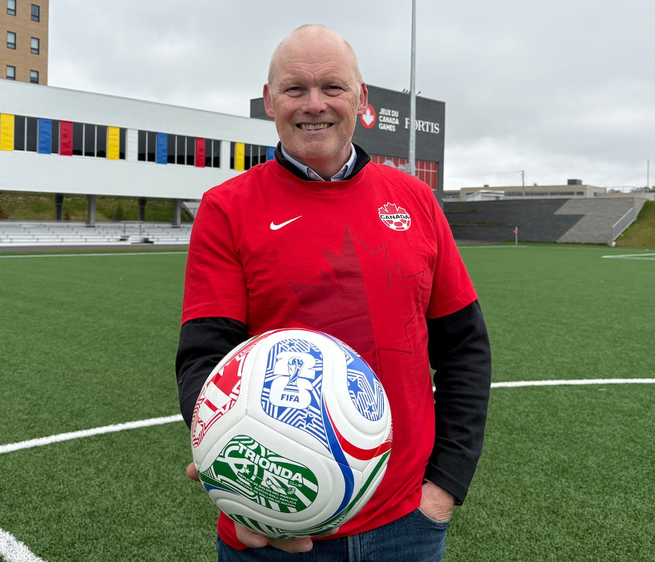 A smiling man wearing a Canada soccer shirt holds the 2026 World Cup ball while standing on a soccer field.