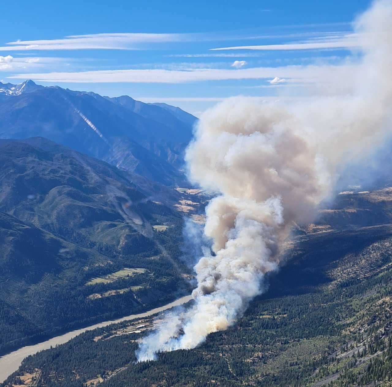 Plumes of smoke arise from a forested area near a river.