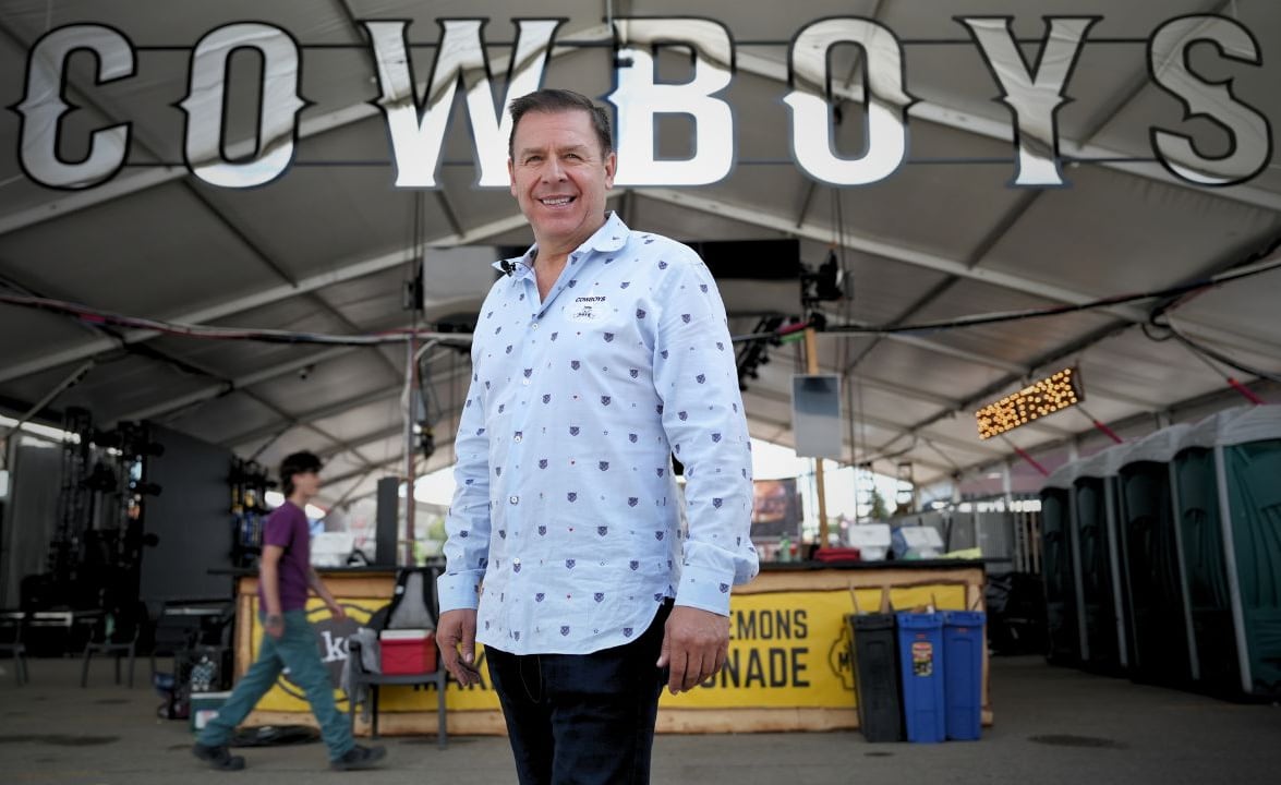 A man stands in front of a banner sign that reads "Cowboys" in silver capital letters hung from a tent with portapotties in the background.