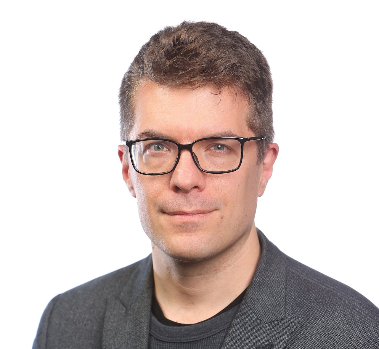 A headshot of a man with short brown hair and glasses, wearing a dark grey blazer over a dark shirt against a white background.