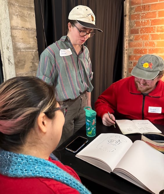 Two people sit at a table, drawing, with another person standing over them.