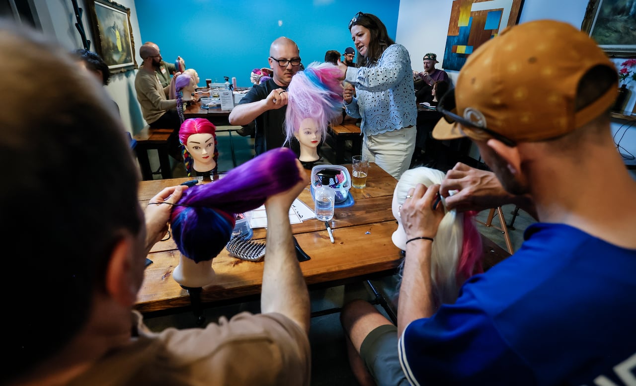 A group of men sit at tables with mannequin heads as they receive help from a woman showing them how to put hair into ponytails, pigtails and braids