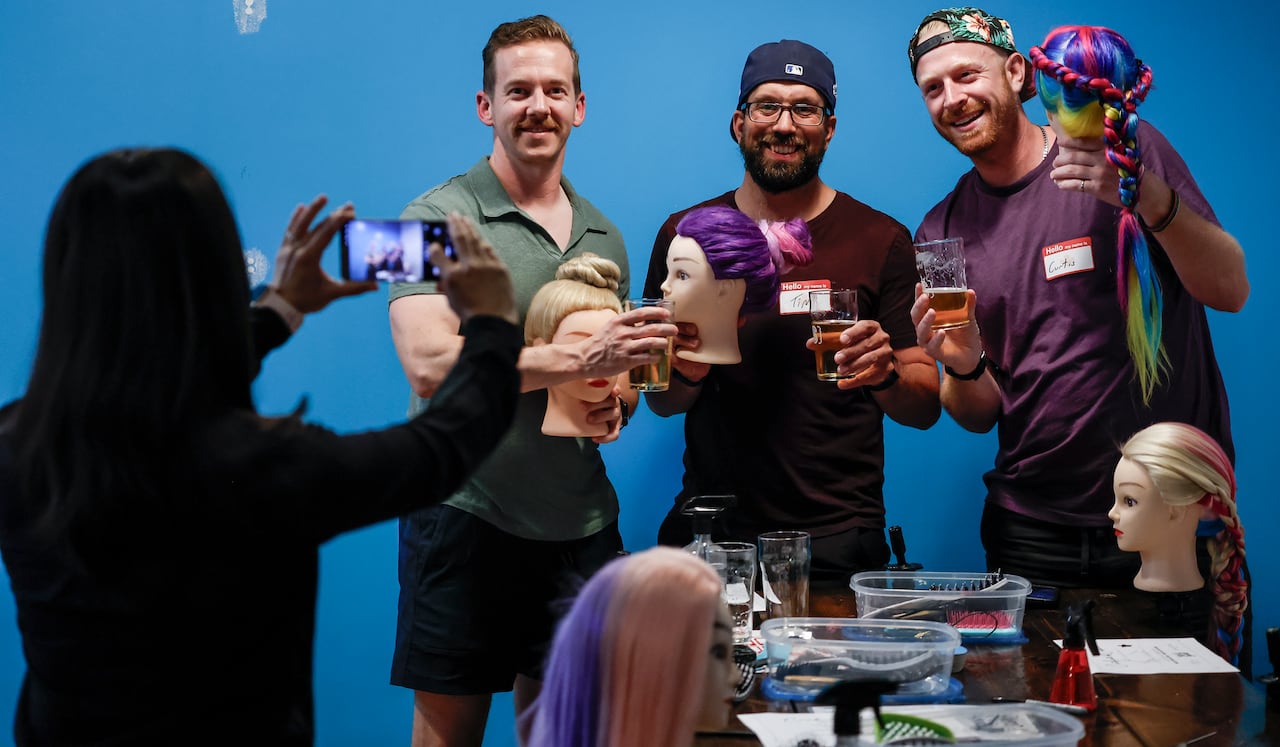 A group of three men smile and hold up mannequin heads with braids and buns while a woman takes a picture on a phone in the foreground.