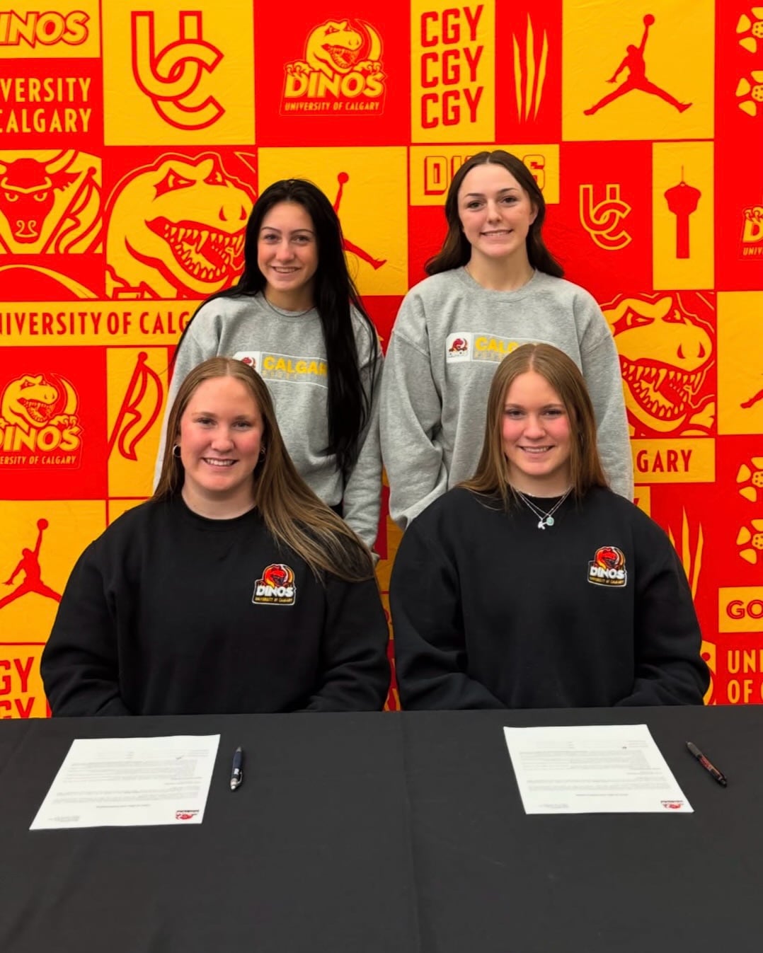Two women sit at a desk with pen and paper in front of them. Two women stand behind them. They are all in front of a University of Calgary Dinos background.
