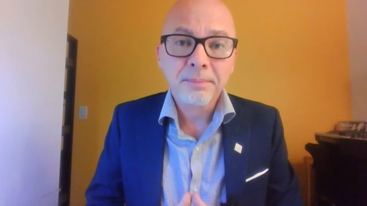 A man in glasses and a blue suit sits in front of a computer camera.