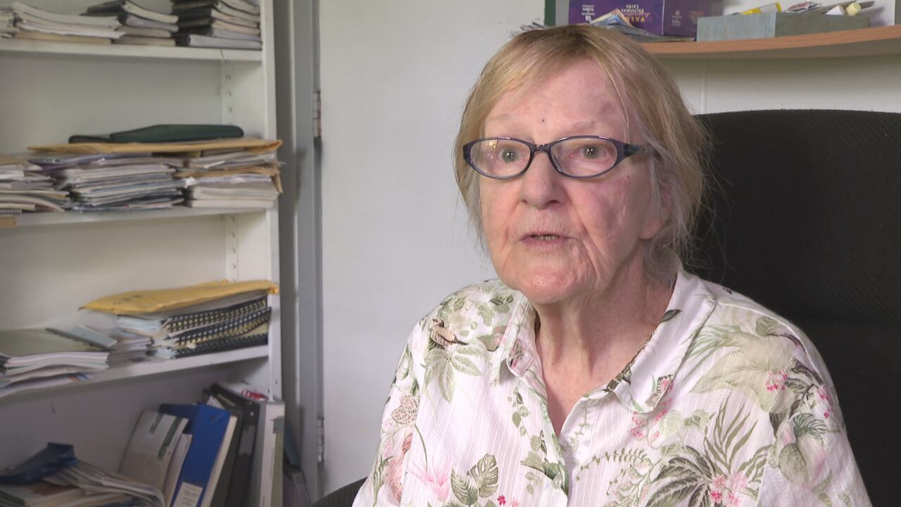 A woman in glasses sits in a chair near a book shelf with many papers.