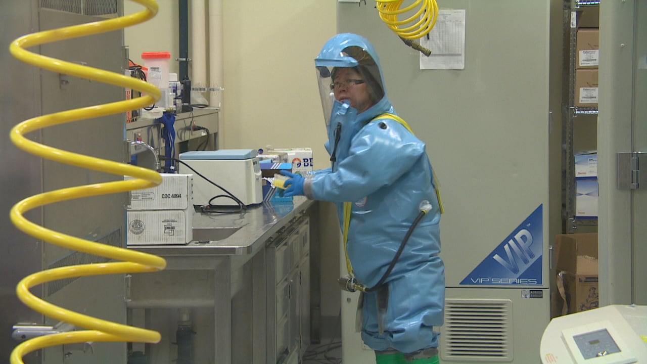 An Asian woman with glasses wears a blue biocontainment suit connected to a respirator while working in a laboratory environment.
