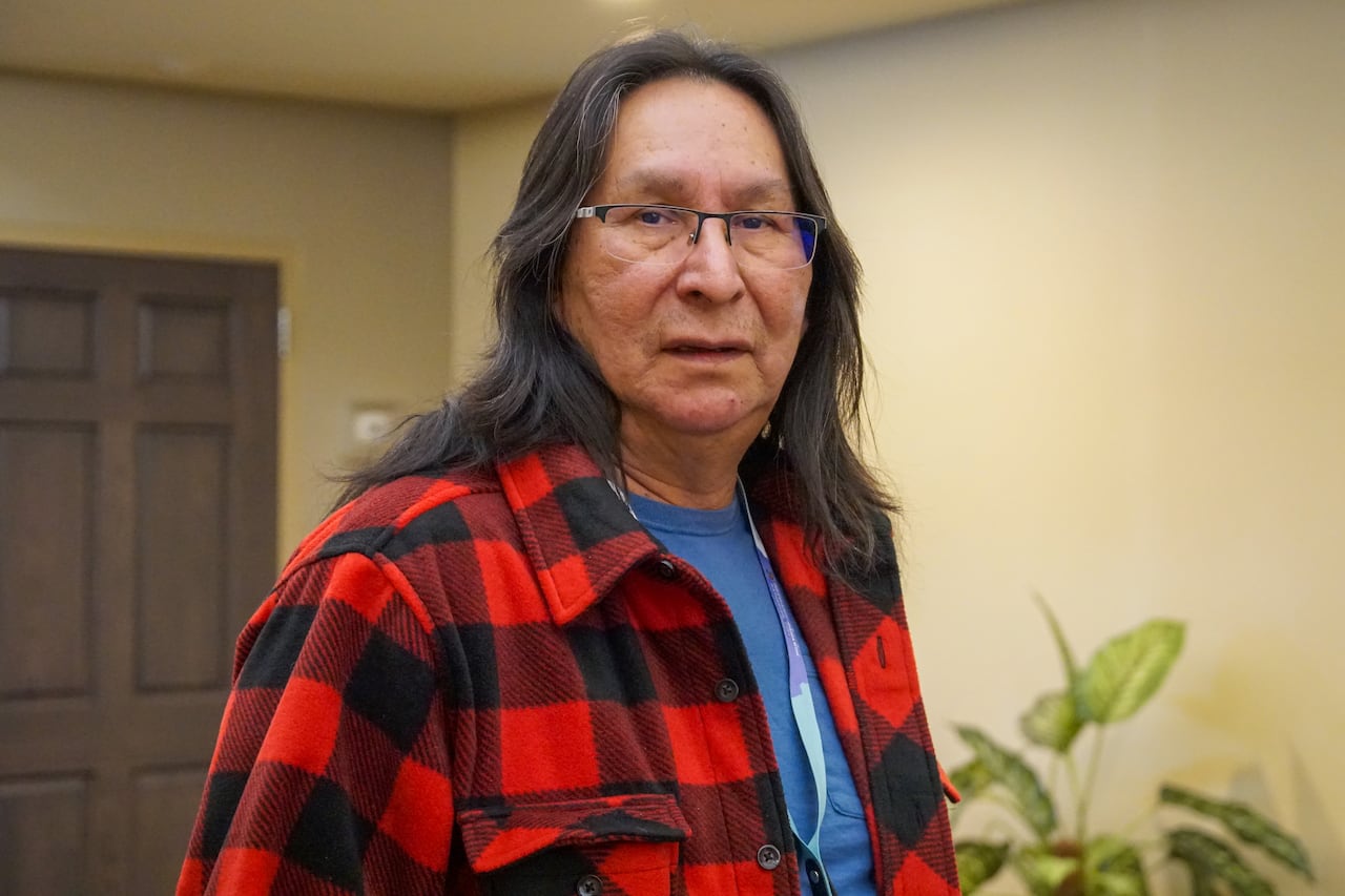 A person with long dark hair and a red plaid shirt stands in a room with a neutral expression on their face.