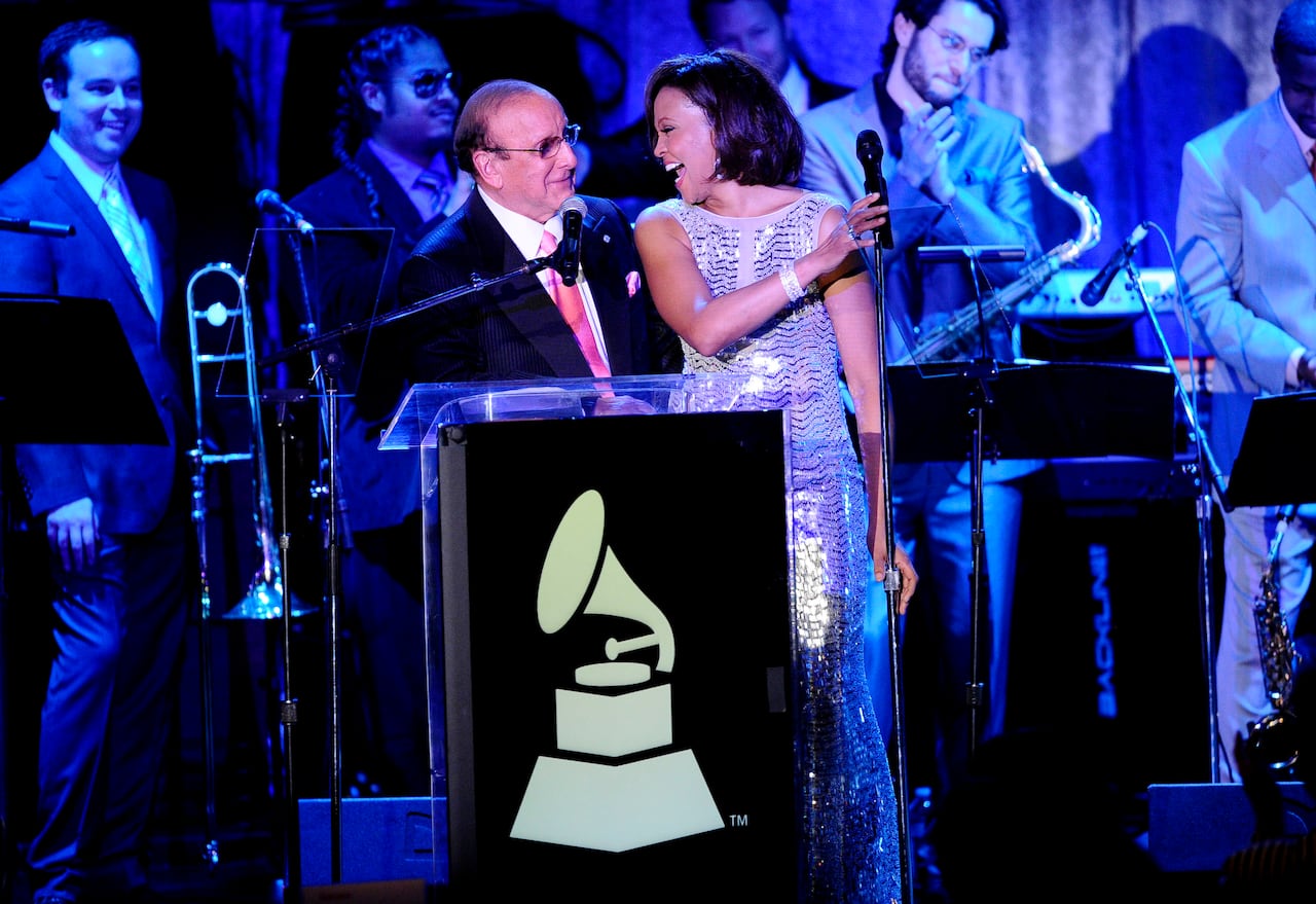 An older, bald  man, wearing glasses, a black suit, white shirt and pink tie, stands next to a Black woman with her hair in a bob cut and wearing a silver gown. The two are smiling at each other on a stage, with musicians in the background.