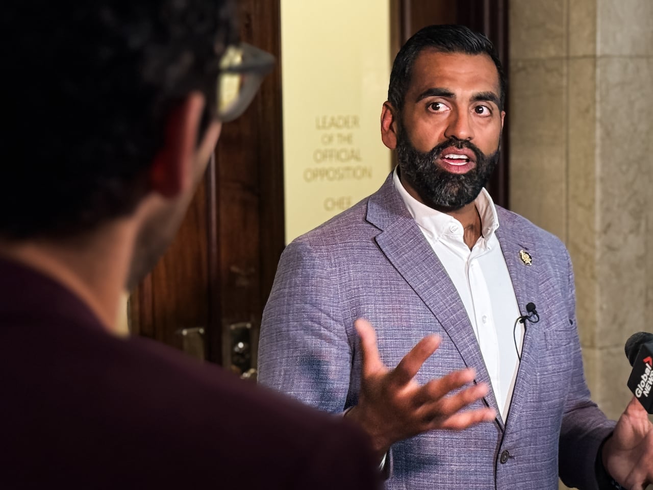 A man with short dark hair and a beard, wearing a light-purple blazer, speaks with reporters outside a legislative office.