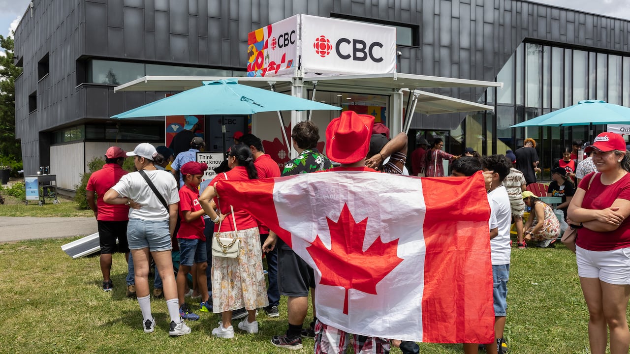 A crowd gathers at an outdoor CBC event on a sunny day. In the center, a person wearing a red cowboy hat holds up a large Canadian flag like a cape.