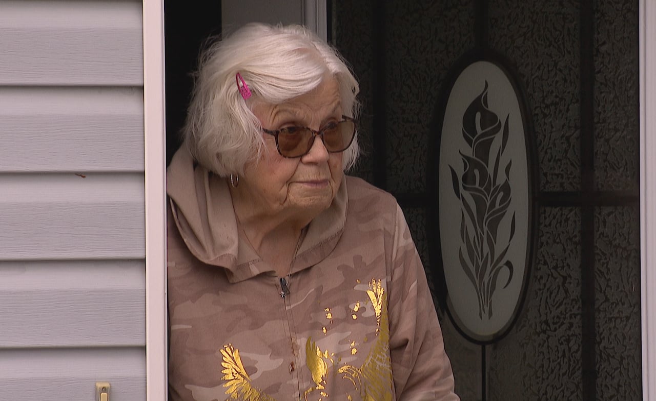 An older woman with white hair stands in front of her door. She wears glasses, a tan raincoat and a pink clip in her hair.