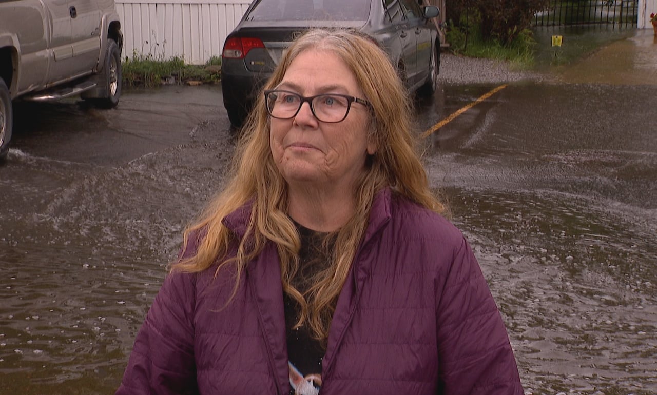 A woman with red hair stands in front of a flooded street, lined with trailer homes. She wears glasses and a purple rain coat.