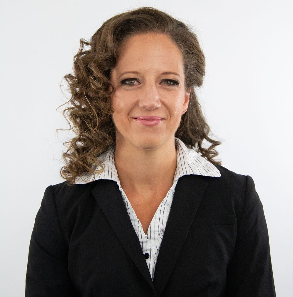 A woman with curly shoulder length brown hair is wearing a black blazer and white blouse
