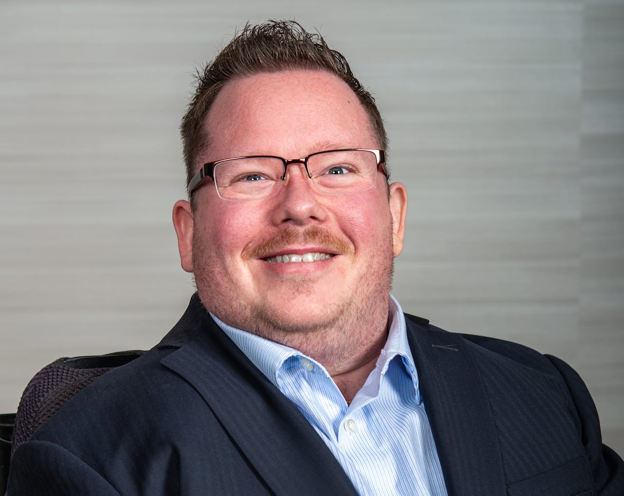 A man wearing glasses is smiling. He's wearing a grey suit jacket and light blue dress shirt