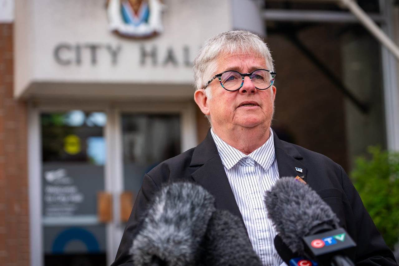 A woman with short white hair speaks in front of a building marked 'City Hall'.