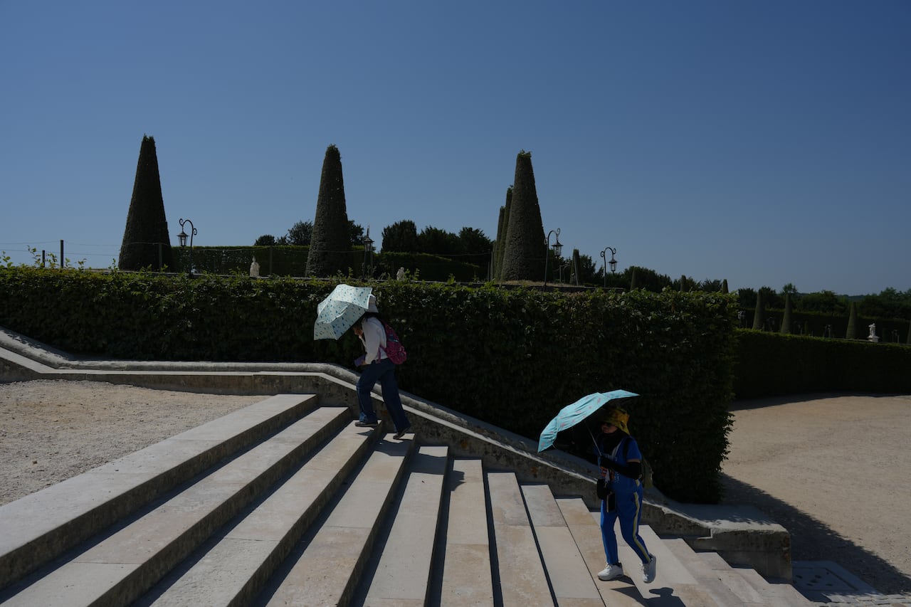 Two people are shown walking up steps from a distance, each with umbrellas, as large shadows are shown on the pavement around them.