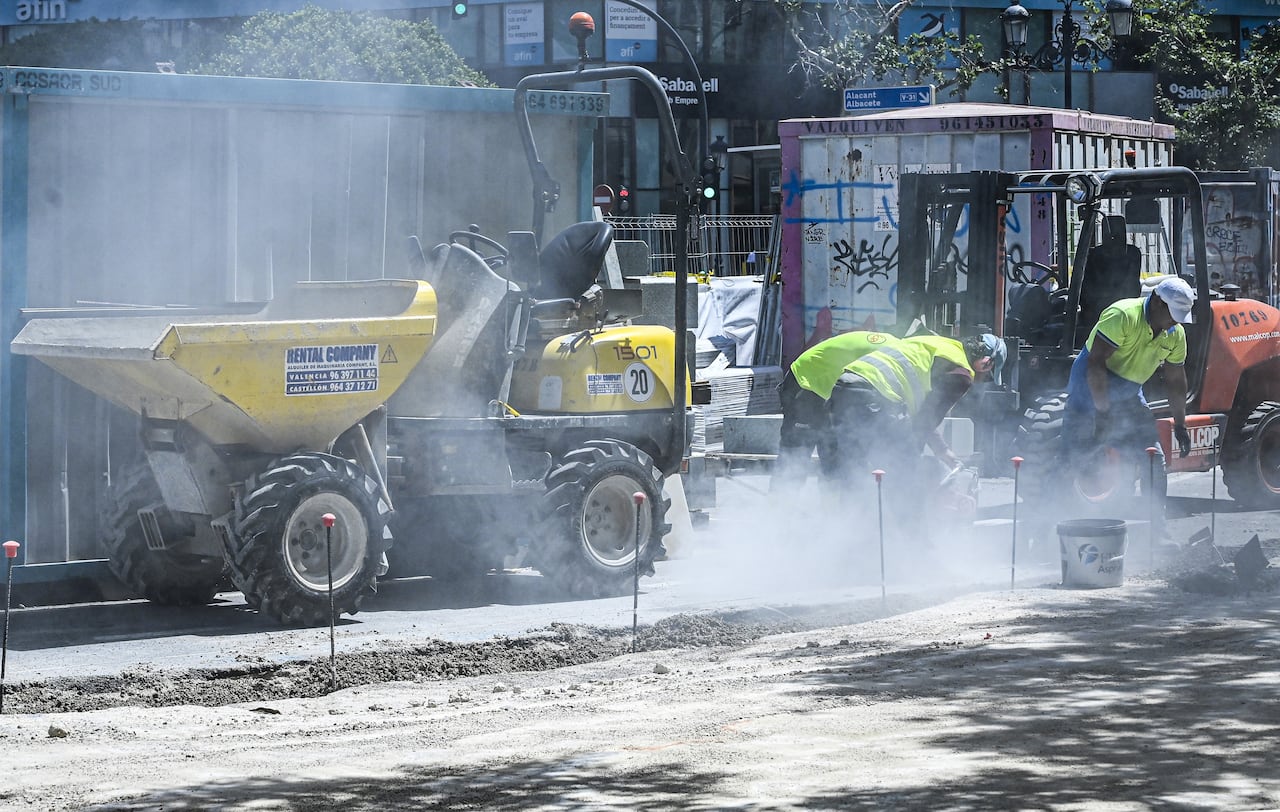 Two construction workers in helmets and reflective clothing are shown working street level near a machine with wheels.