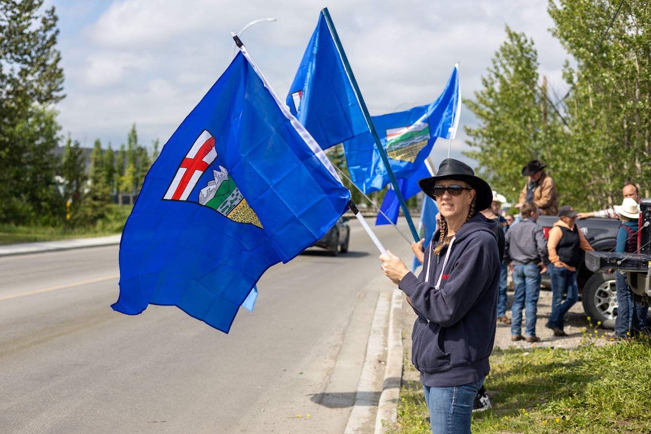 A row of people hold Alberta flags on the side of a street. A woman wearing a cowboy hat is standing nearest the camera, flying a flag.