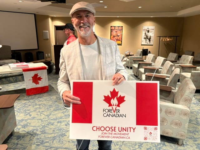 A man in a hat holds a lawn sign with a Canadian flag on it.