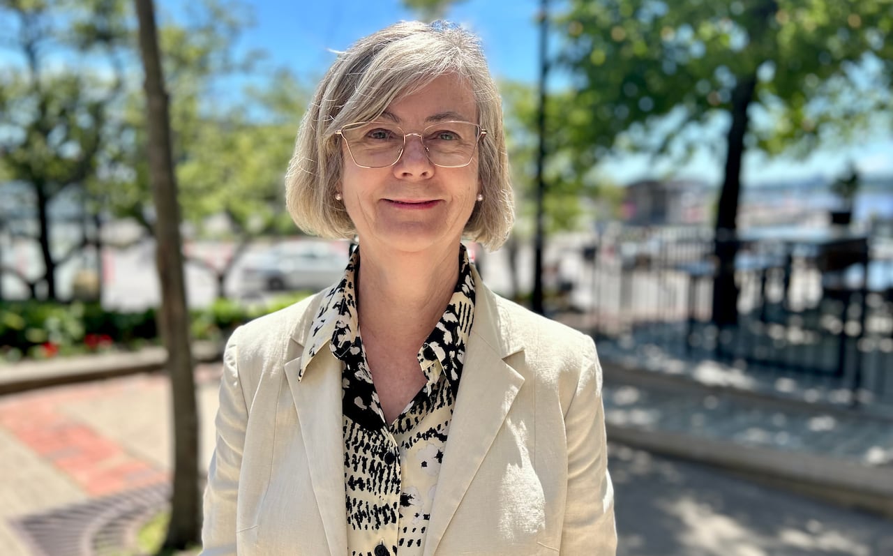 A woman with a tan blazer smiles at the camera while standing outside in summer with city infrastructure behind her.