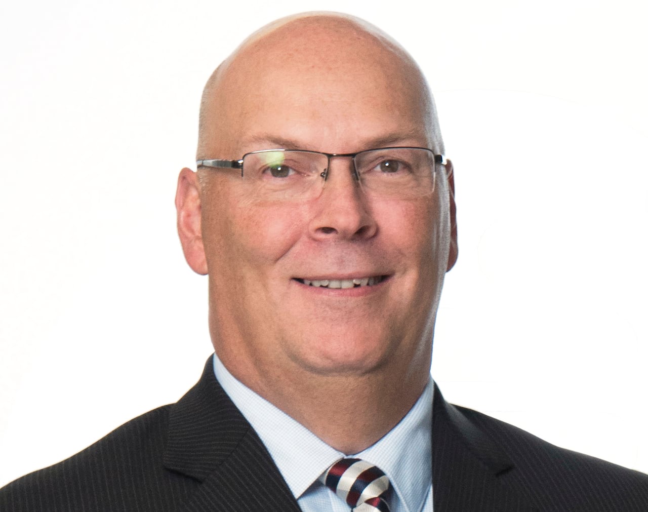 A man with a black suit, tie and glasses, smiles at the camera with a white background.